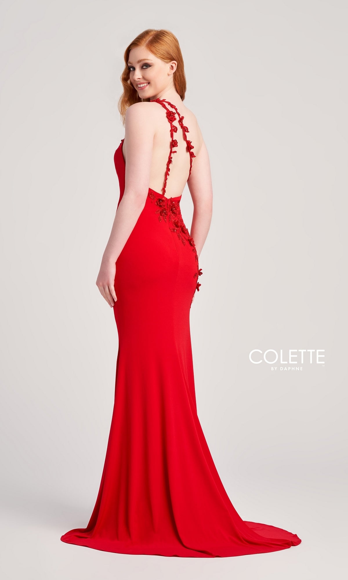 A woman wears the Colette One-Shoulder Sleek Long Prom Dress CL5108, a fitted red gown with floral details and an open back, standing against a plain backdrop and gracefully looking over her shoulder.